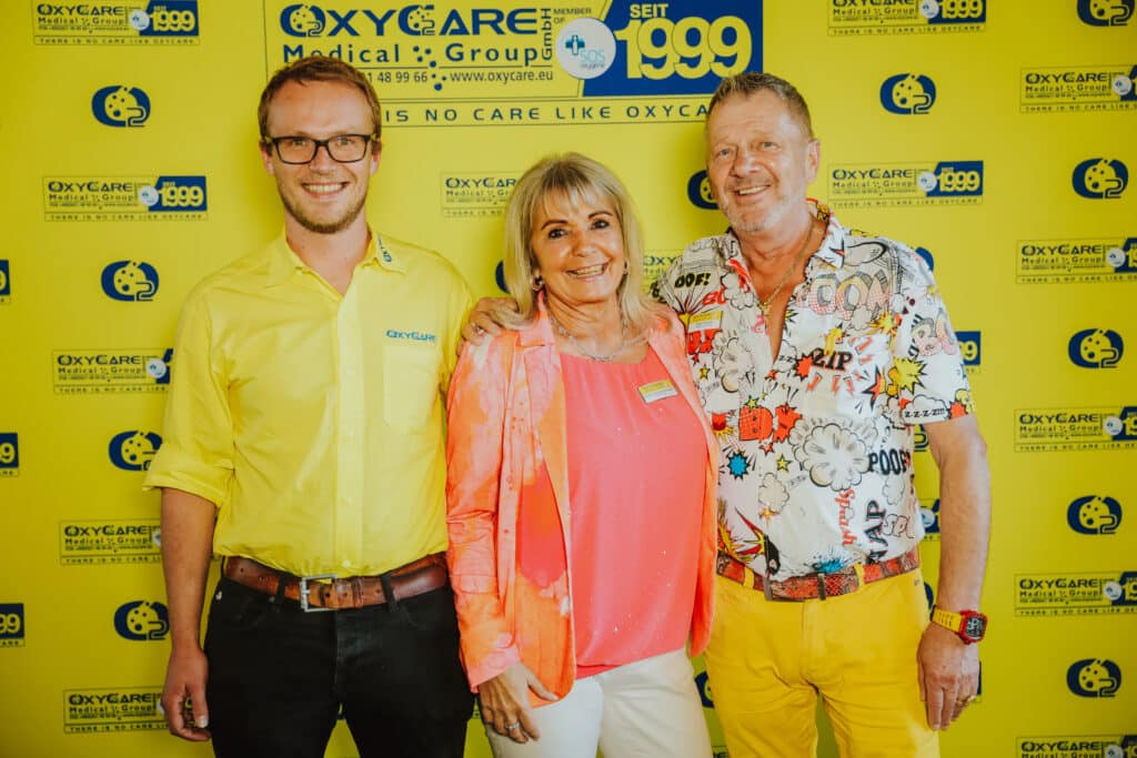 Auf dem Foto stehen drei Personen vor einer OxyCare Fotowand. Von links nach rechts: Pierre Lange, Andrea Schardey-Peschel und Peter Peschel. Alle lächeln und stehen eng beieinander.